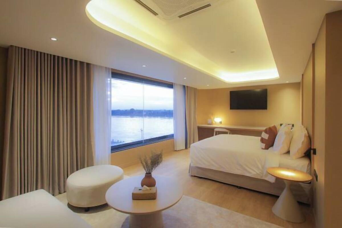 A modern hotel room with a large bed, a desk, a chair, and a view of the water.