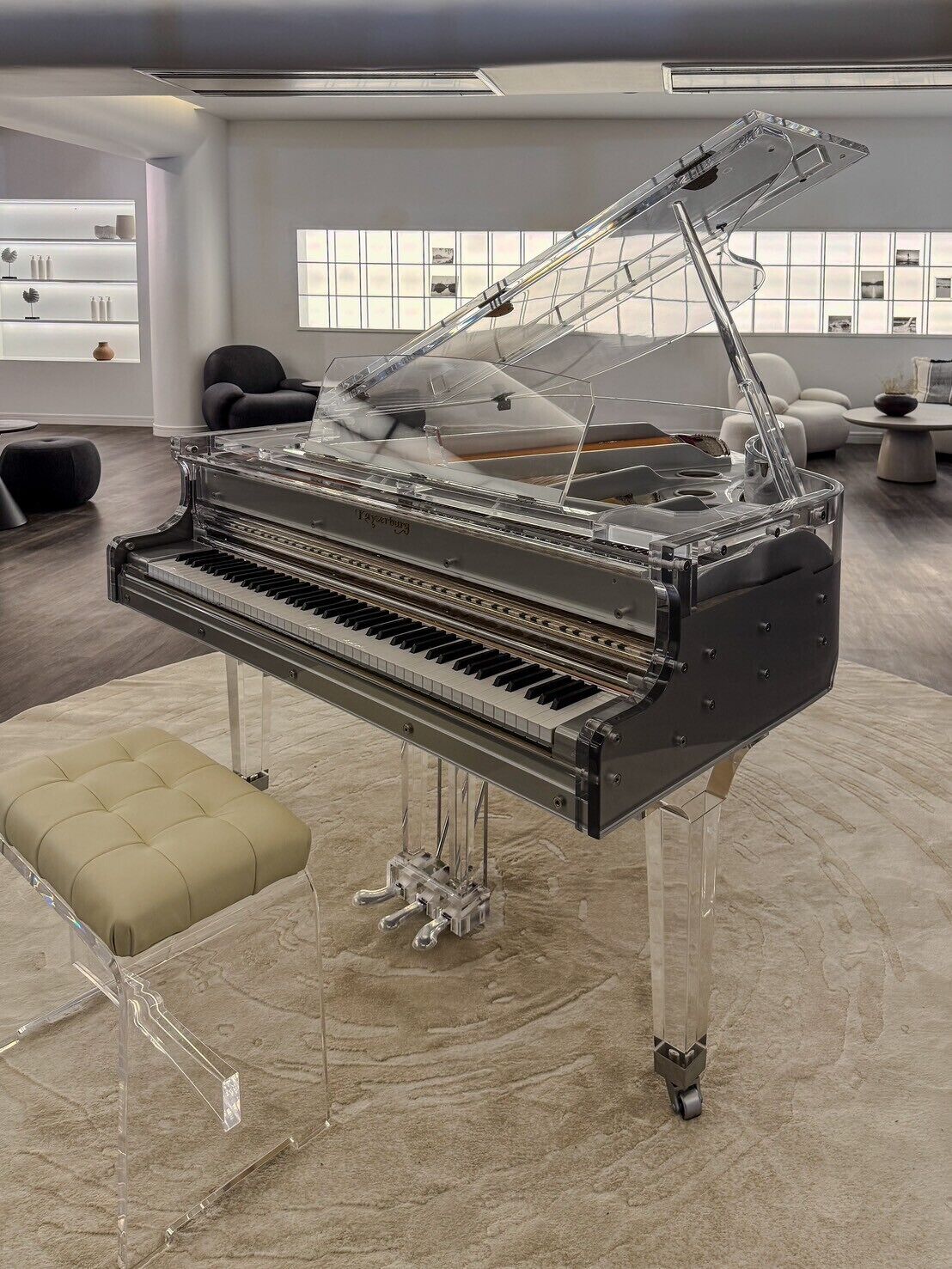 A grand piano with a glass top and legs, placed in a modern room with a large window, a sofa, and a coffee table.