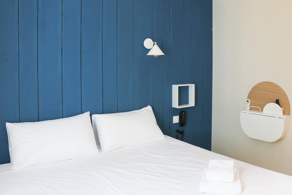 A neatly made bed with white linens, a wall-mounted lamp, a phone, and a small wooden shelf with a mirror and a towel.