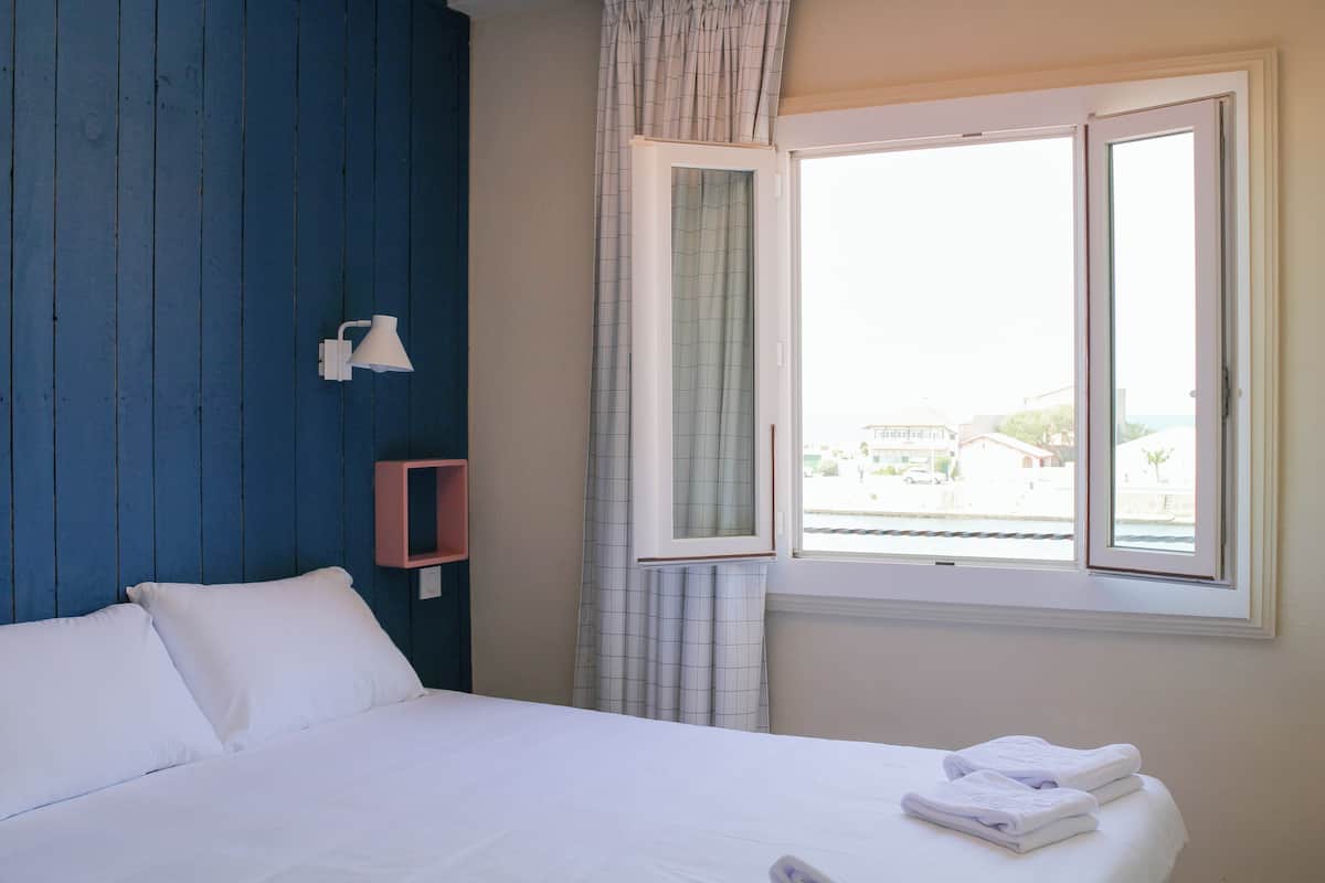 A bedroom with a bed, a window with curtains, a wall-mounted lamp, and a small pink shelf.