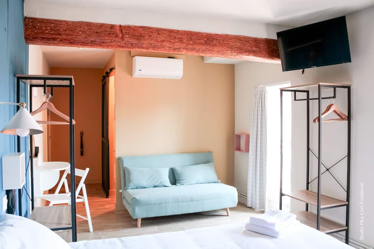 A modern hotel room with a light blue sofa, a TV mounted on the wall, a wooden ceiling, and a clothing rack.
