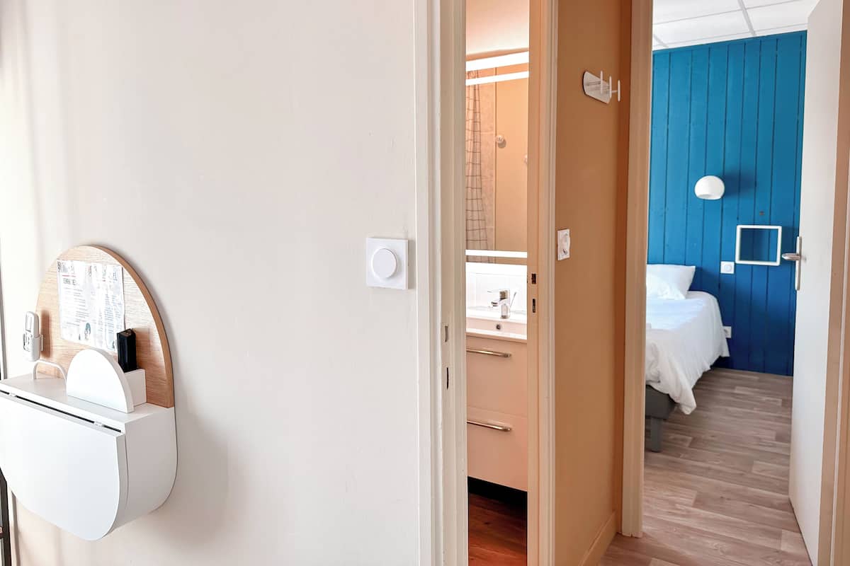 A hotel room with a wooden floor, a bed with white bedding, a blue accent wall, and a small white-framed mirror.
