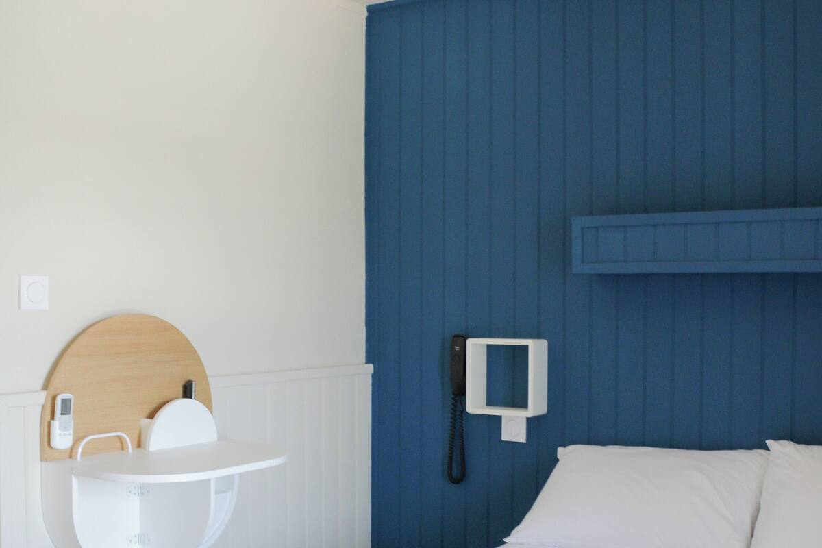 A hotel room with a bed, a bedside table, a wall-mounted phone, and a small shelf with a mirror and a phone.