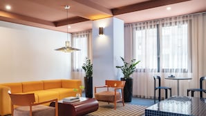 A modern living room with a yellow sofa, two wooden chairs, a coffee table, and a small round table with chairs. There are two potted plants, a patterned rug, and a ceiling light. - Numa Rome Trastevere (Rome)