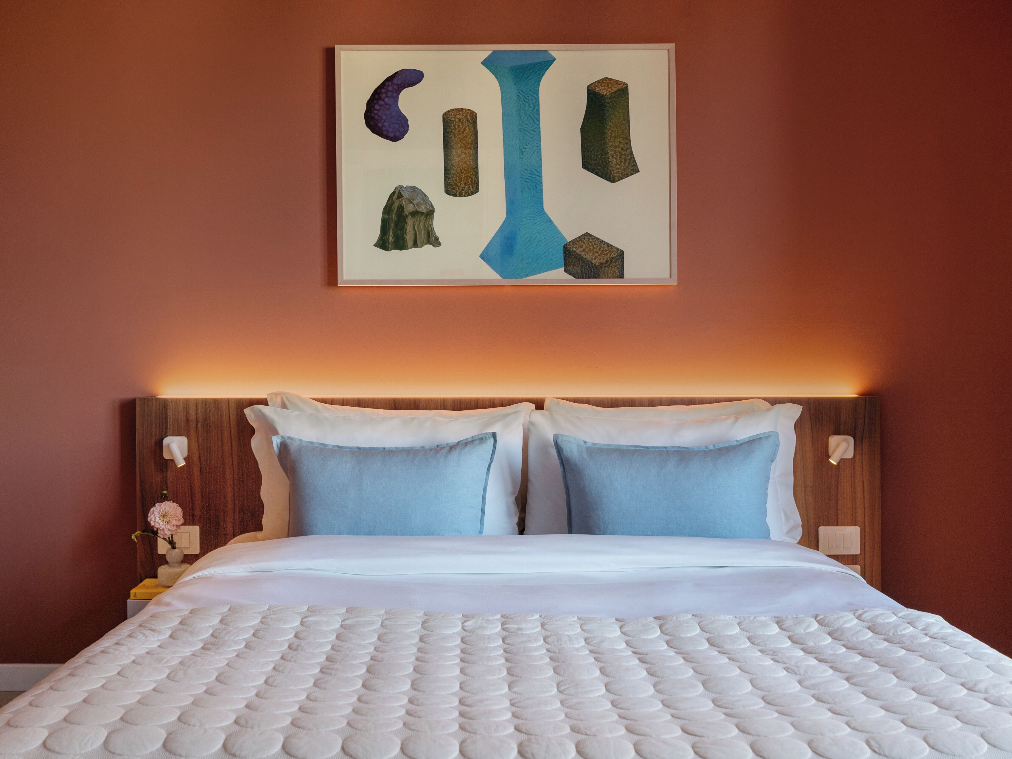 Ein ordentlich bezogenes Bett mit weißer Daunendecke und zwei blauen Kissen, ein Holz-Kopfteil und ein gerahmtes abstraktes Kunstwerk darüber.