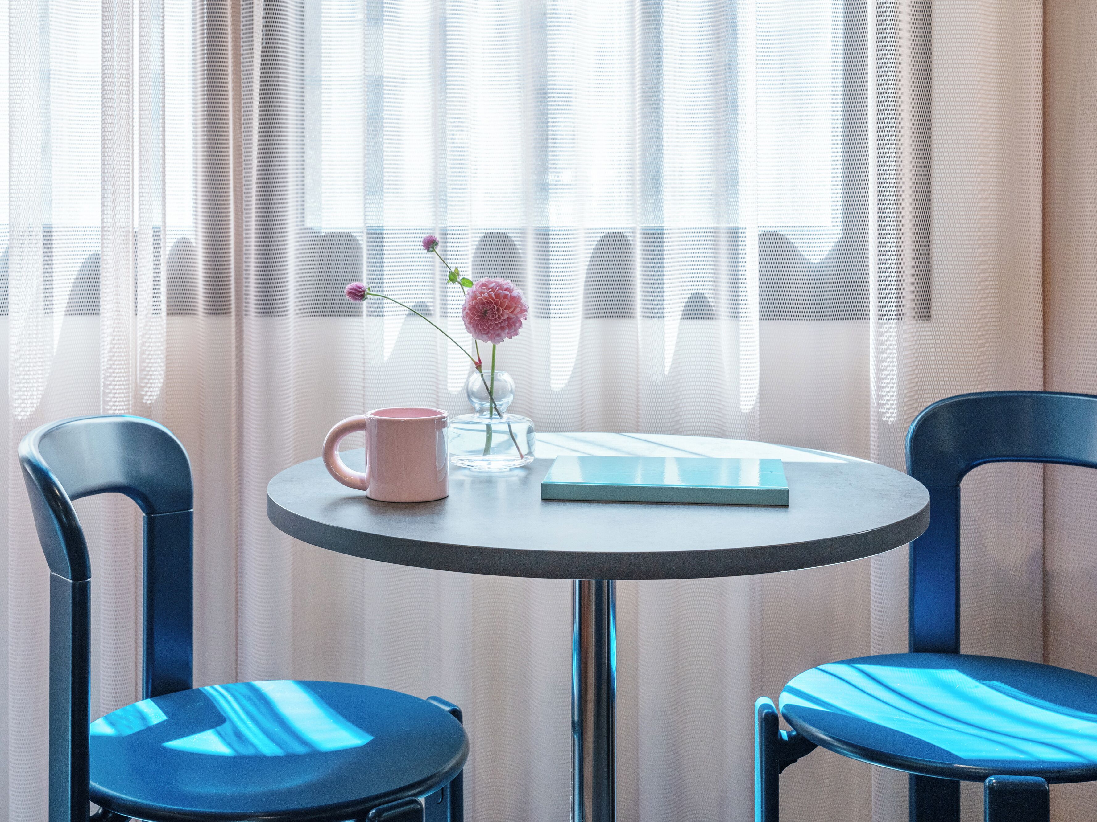 A round table with a pink mug, a book, and a vase with a flower. Two blue chairs are positioned around the table.