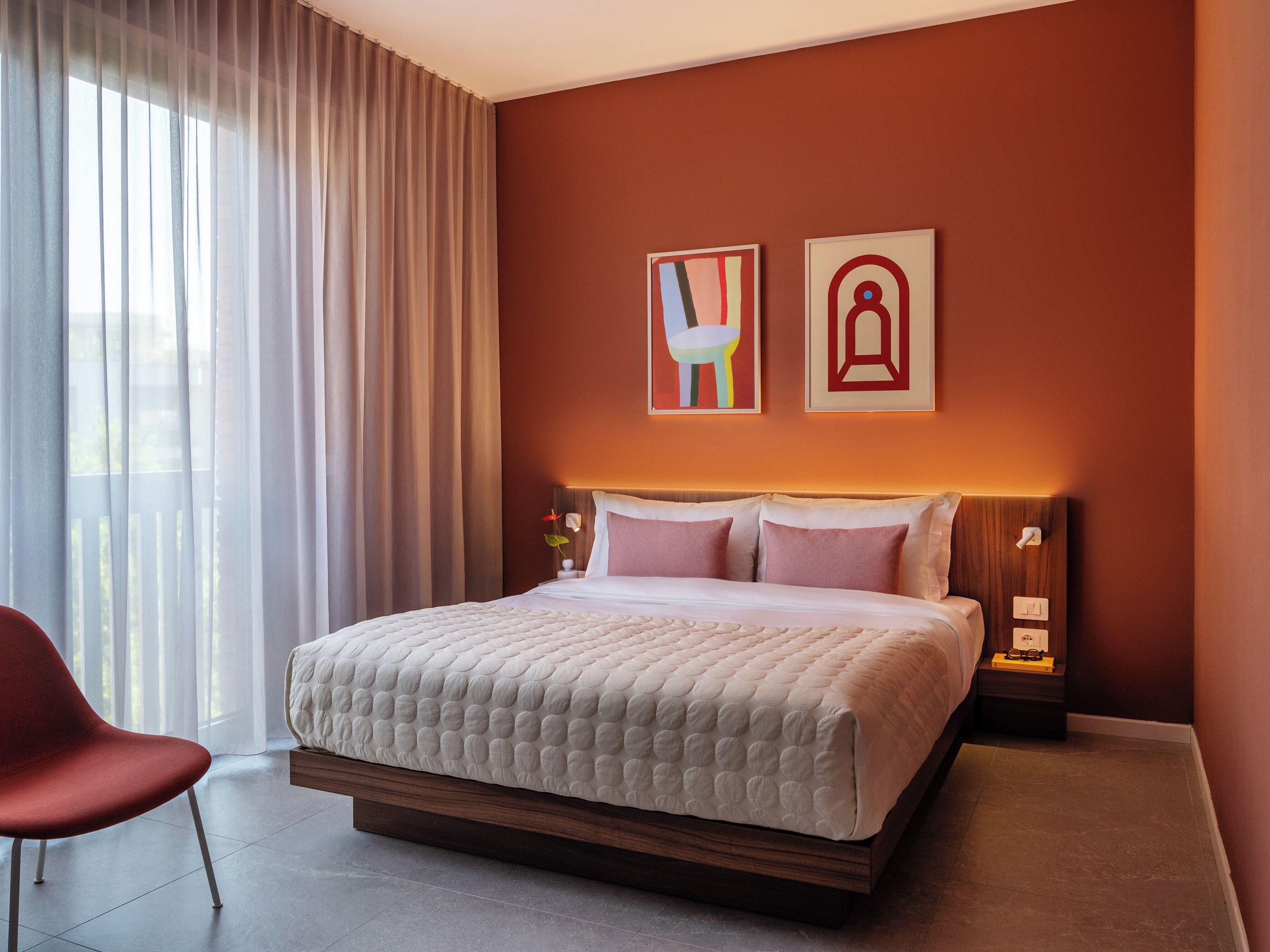 A hotel room with a large bed, a red chair, and two wall-mounted artworks.
