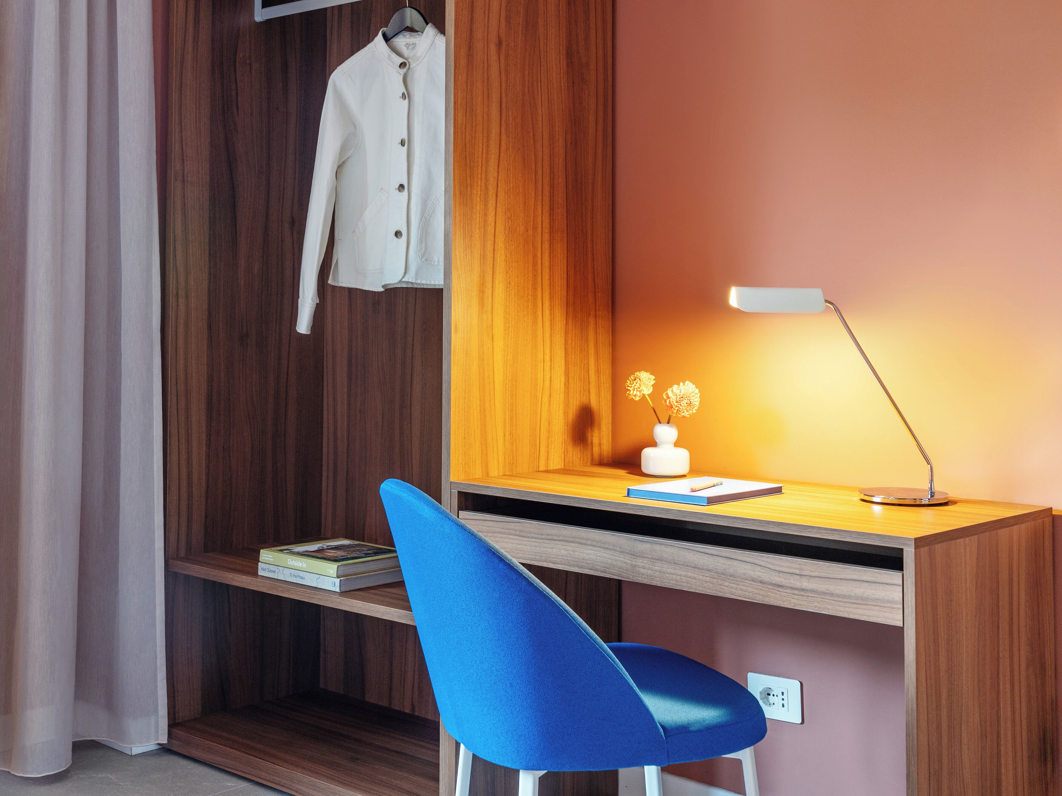 A modern workspace with a wooden desk, a blue chair, a white vase with flowers, and a hanging coat.