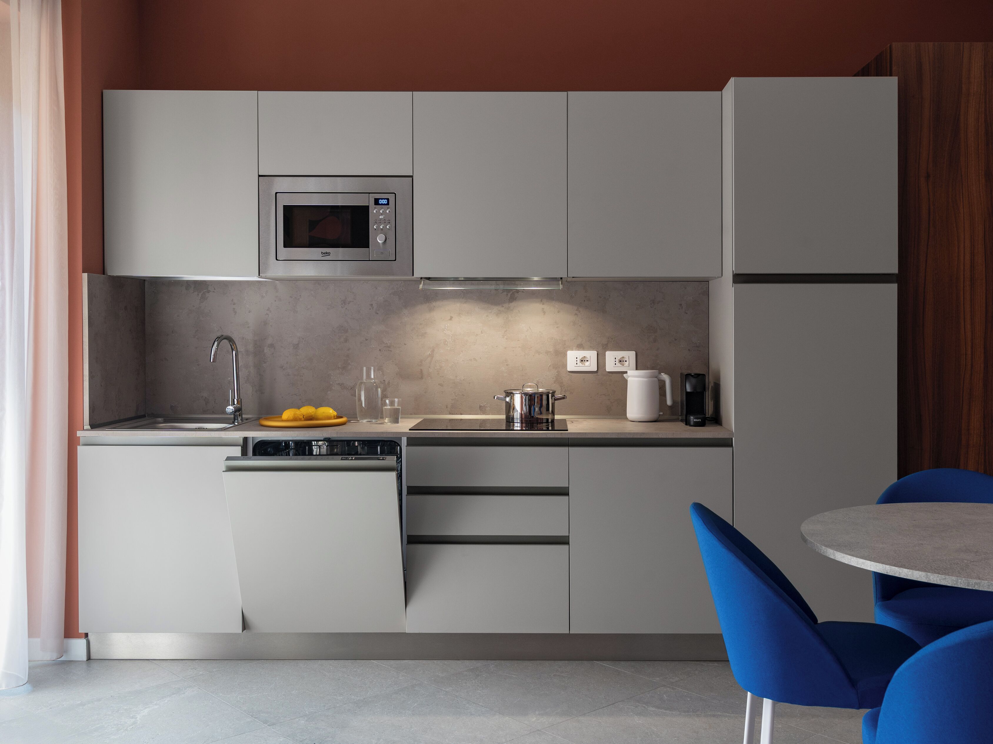 Eine moderne Küche mit integriertem Mikrowellenherd, Spüle und Kaffeemaschine. Ein Essbereich mit blauen Stühlen und einem kleinen Tisch.