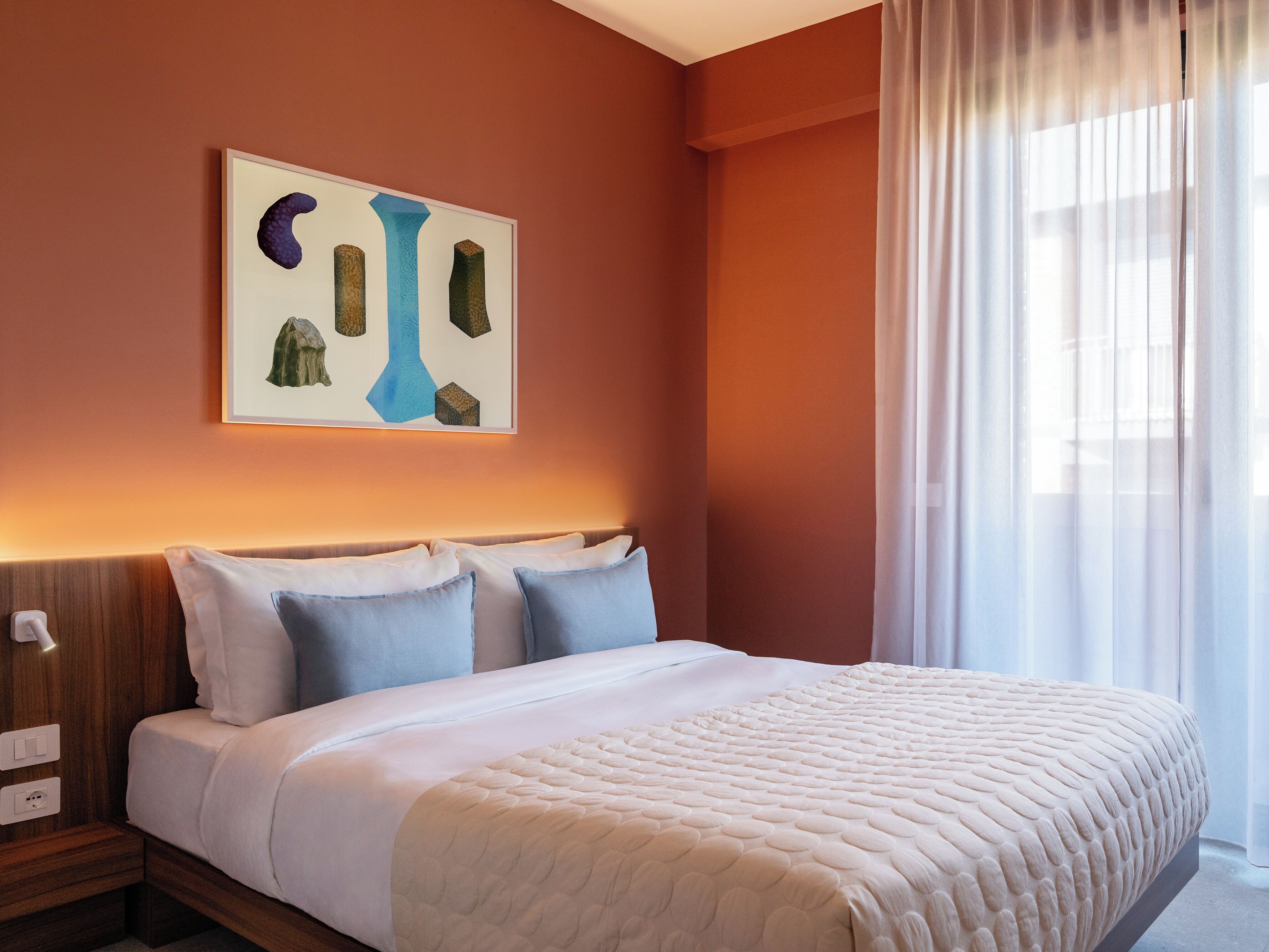 A hotel room with a bed, a nightstand, a wall-mounted artwork, and a window with curtains.