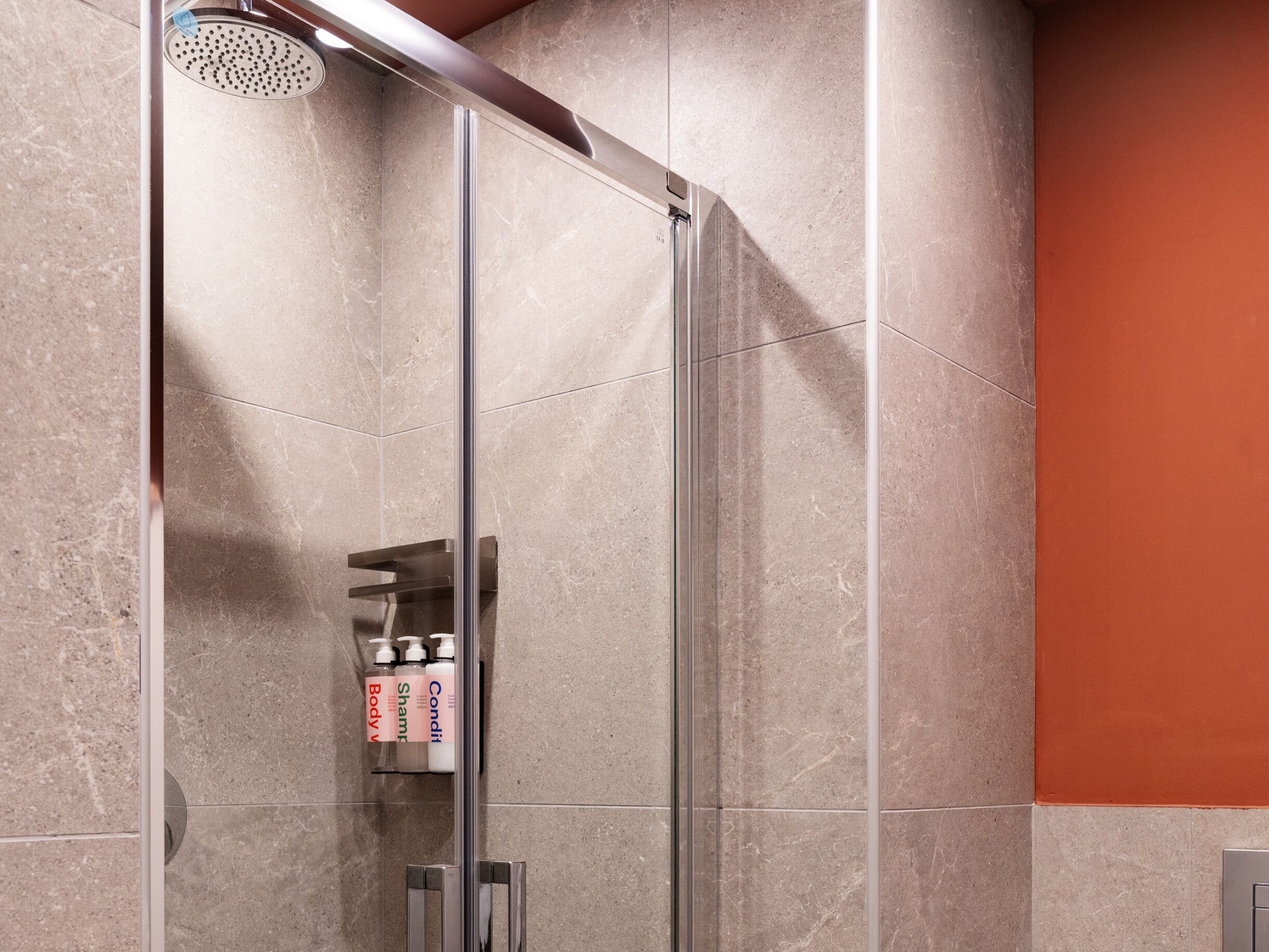 A glass-enclosed shower stall with a handheld showerhead, a shelf with toiletries, and a tiled wall.