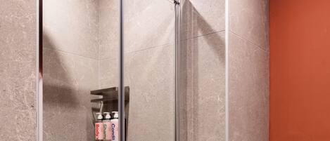 A glass-enclosed shower stall with a handheld showerhead, a shelf with toiletries, and a tiled wall.