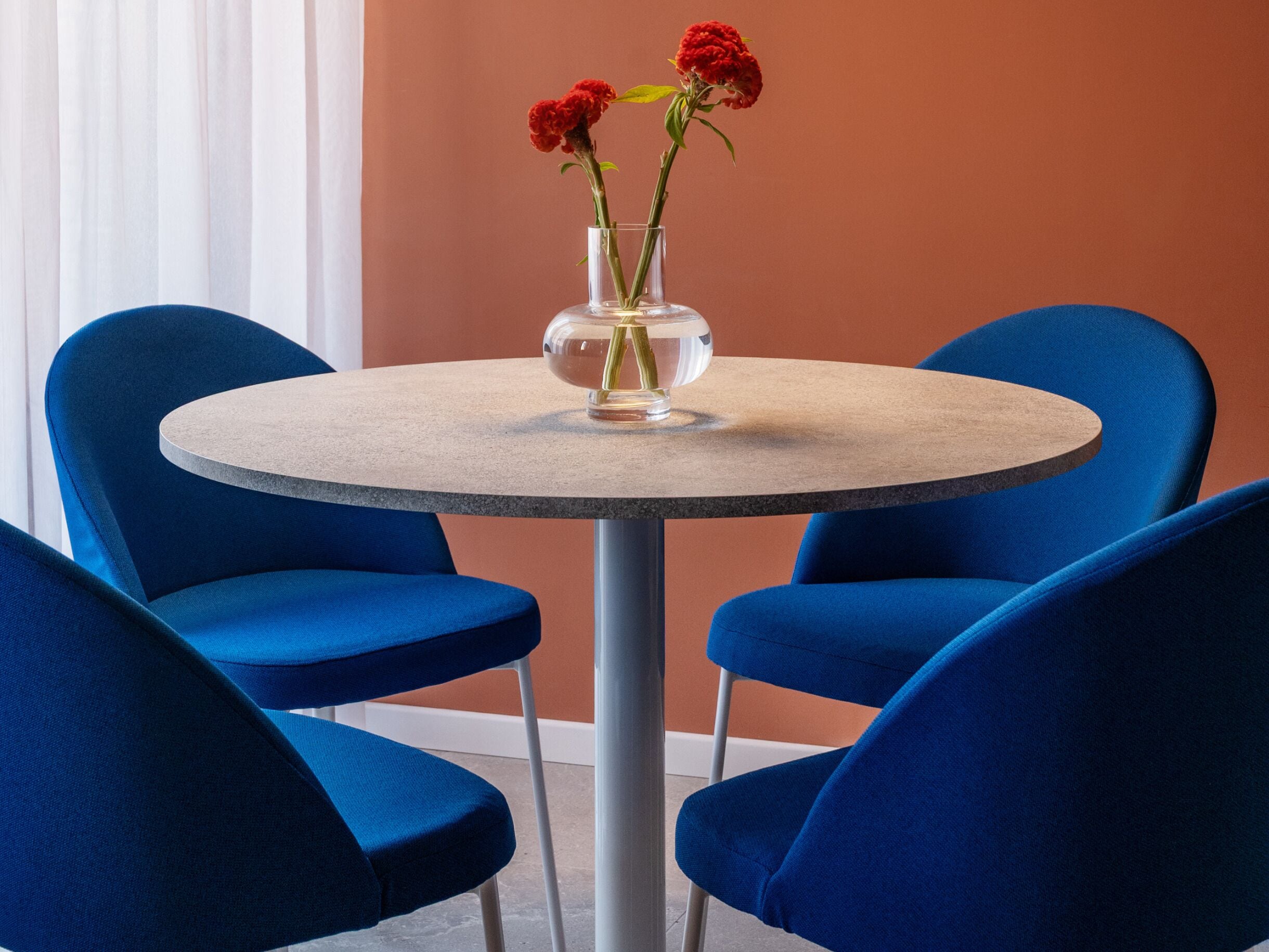 A round table with a vase of red flowers, surrounded by four blue chairs.