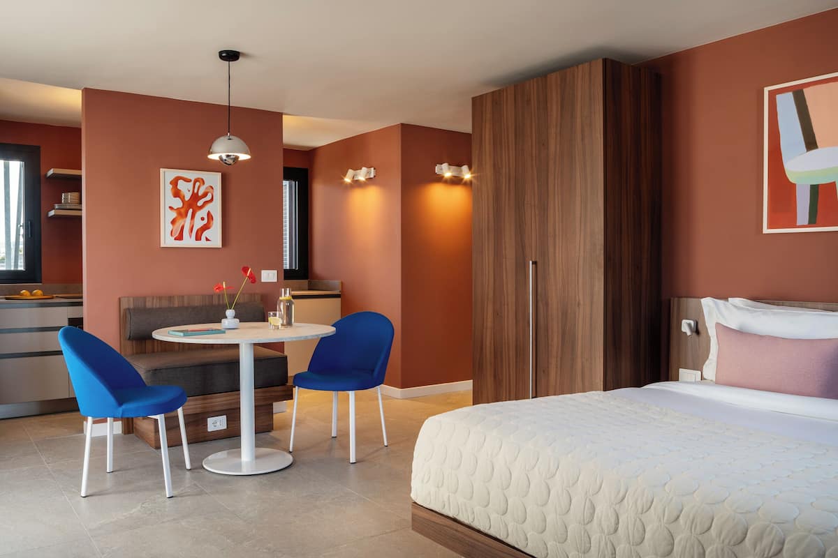 Una habitación de hotel moderna con una cama, dos sillas azules, una mesa redonda, un armario de madera y una kitchenette al fondo.