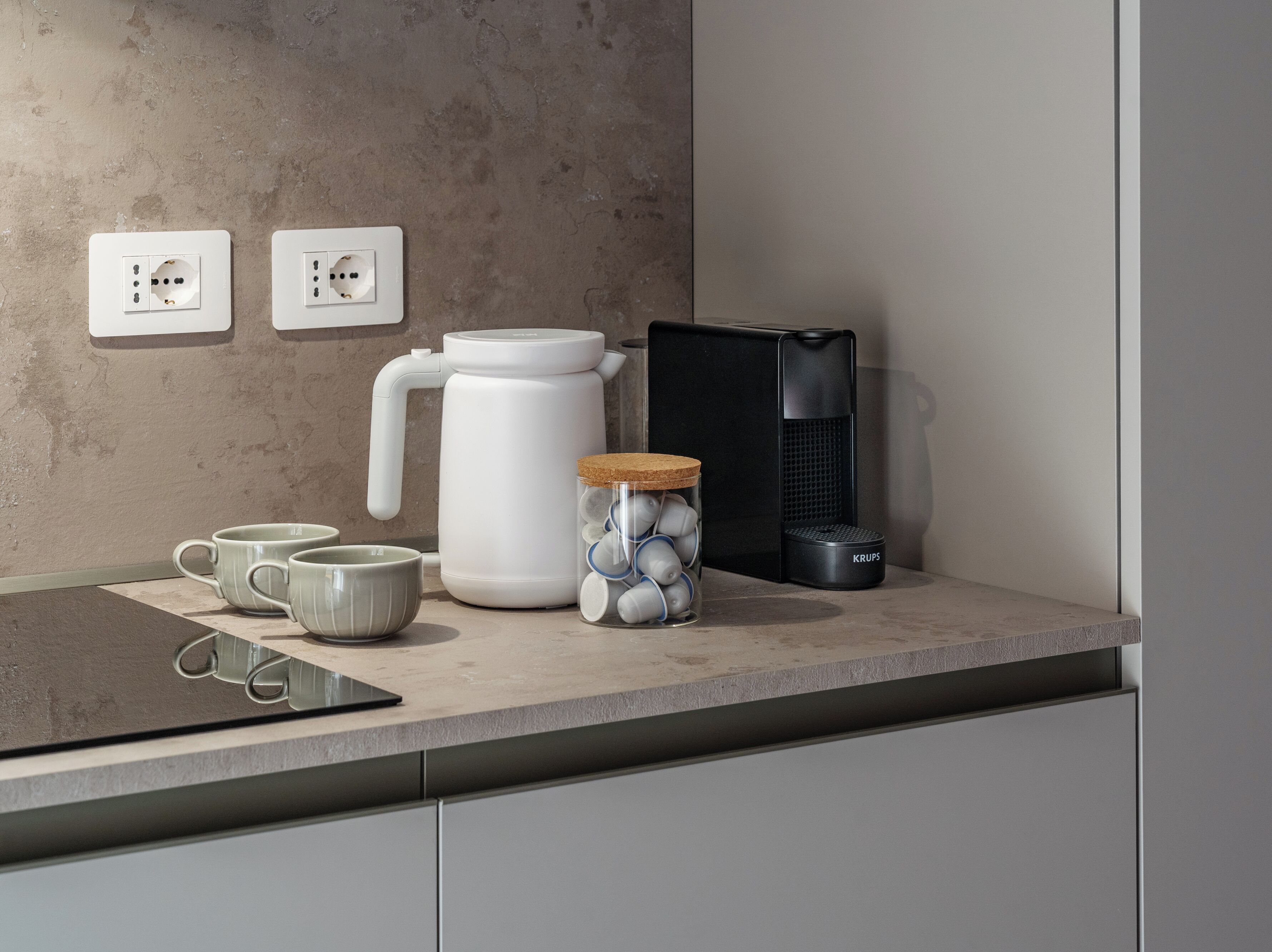 A kitchen counter with a coffee maker, a kettle, a jar with tea bags, and a black appliance.