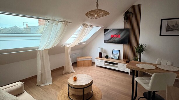 A modern living room with a sofa, a round coffee table, a dining table with chairs, a TV mounted on the wall, and a view of the city through large windows. - Cathedral view apartment (Bayeux)