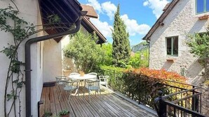 A wooden deck with a table and chairs, surrounded by greenery and a stone building.