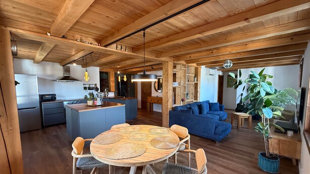A spacious living area with a wooden ceiling, a blue sofa, a dining table with woven placemats, and a kitchen with modern appliances.