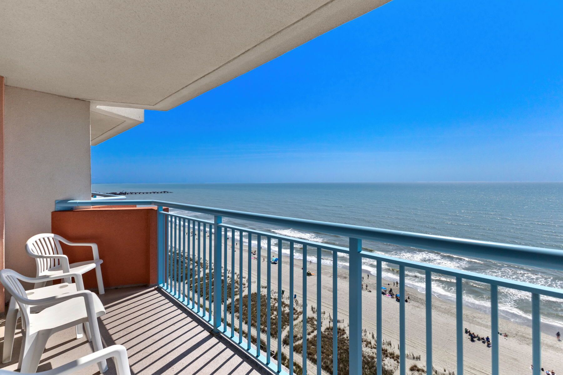 A balcony with two white plastic chairs, a view of the beach, and a clear blue sky.