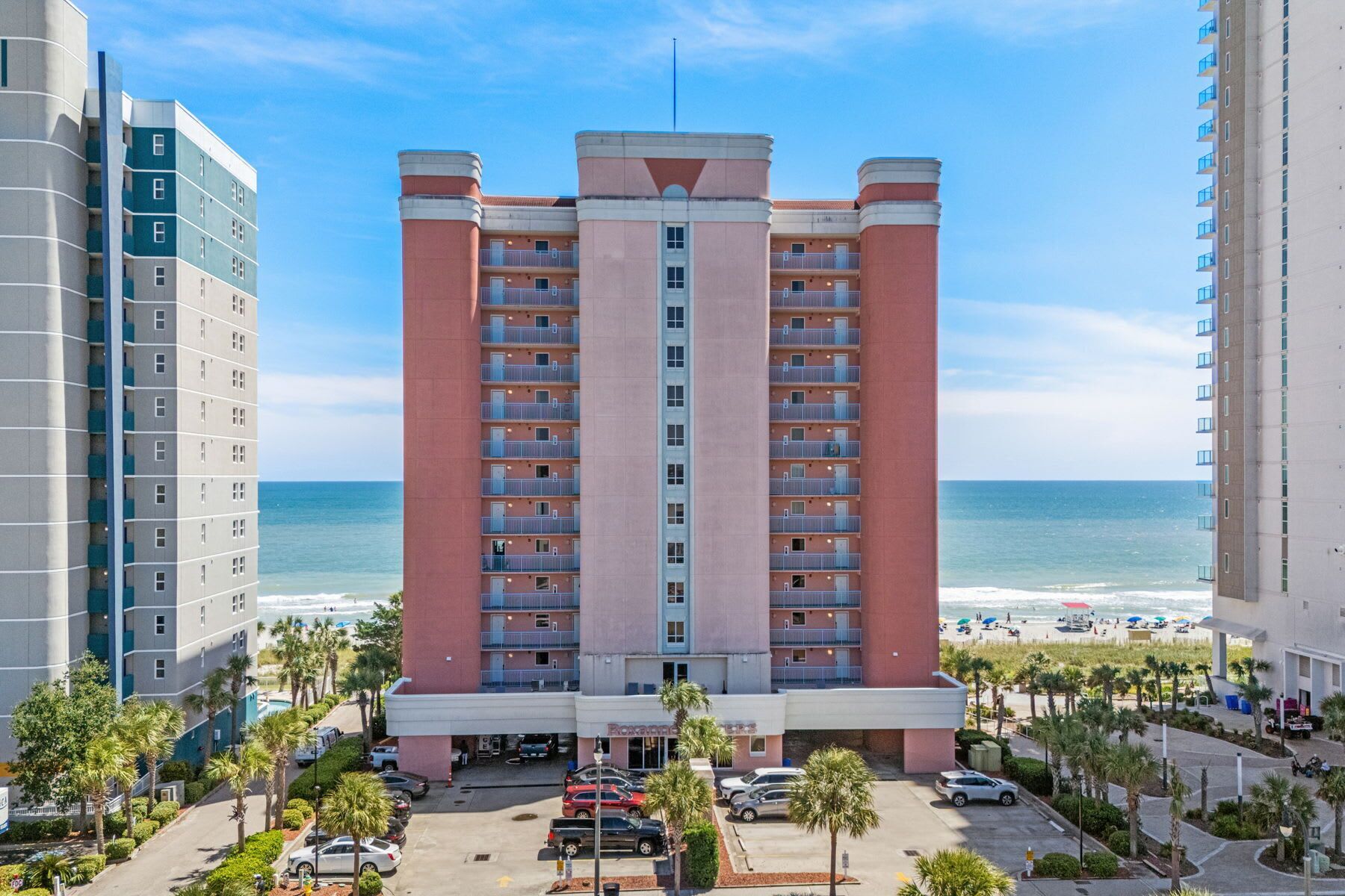 A high-rise hotel with multiple balconies, a parking lot, and a beach view.