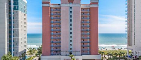 A high-rise hotel with multiple balconies, a parking lot, and a beach view.
