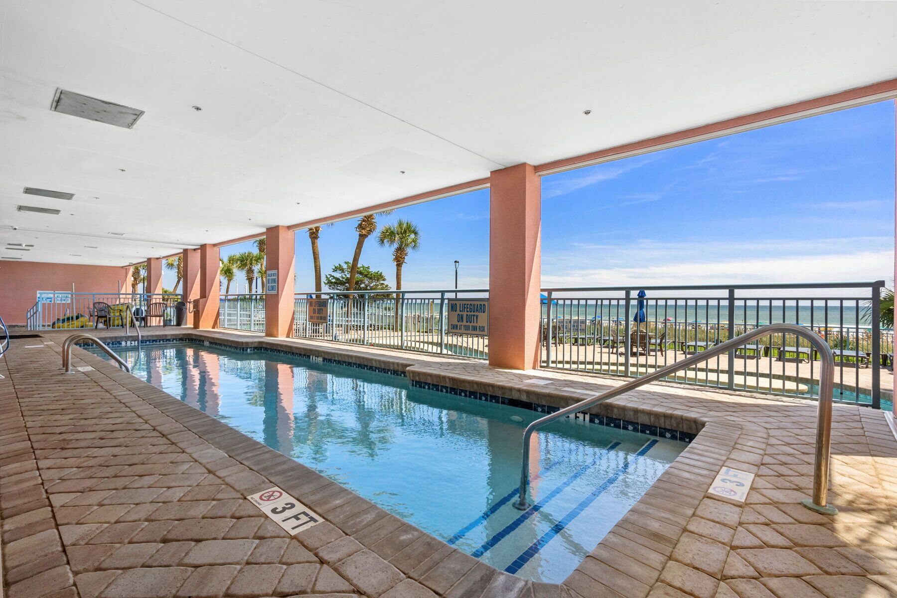 Une piscine dont la profondeur est indiquée à 3 pieds, entourée d’une terrasse avec une rambarde, de palmiers et offrant une vue sur la plage.