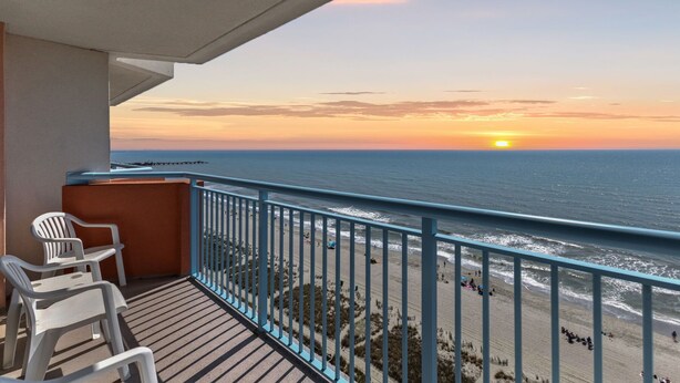 A balcony with a view of the ocean at sunset, featuring a railing and two white chairs.