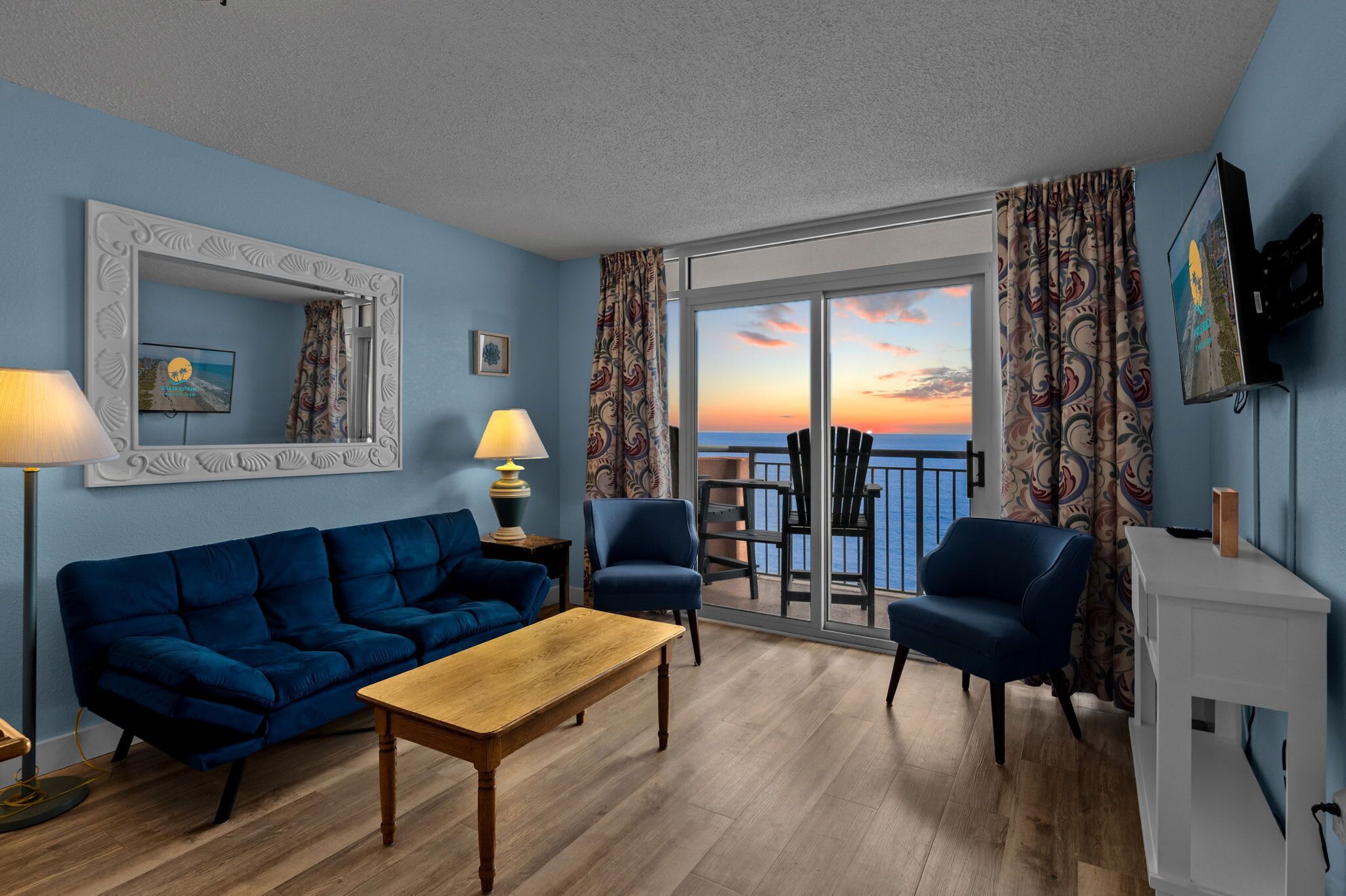 A living room with a blue sofa, a wooden coffee table, and two armchairs. There is a large window with a view of the sunset and the sea.