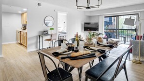 A modern dining area with a wooden table set for four, a chandelier, and a view of the city through large windows.