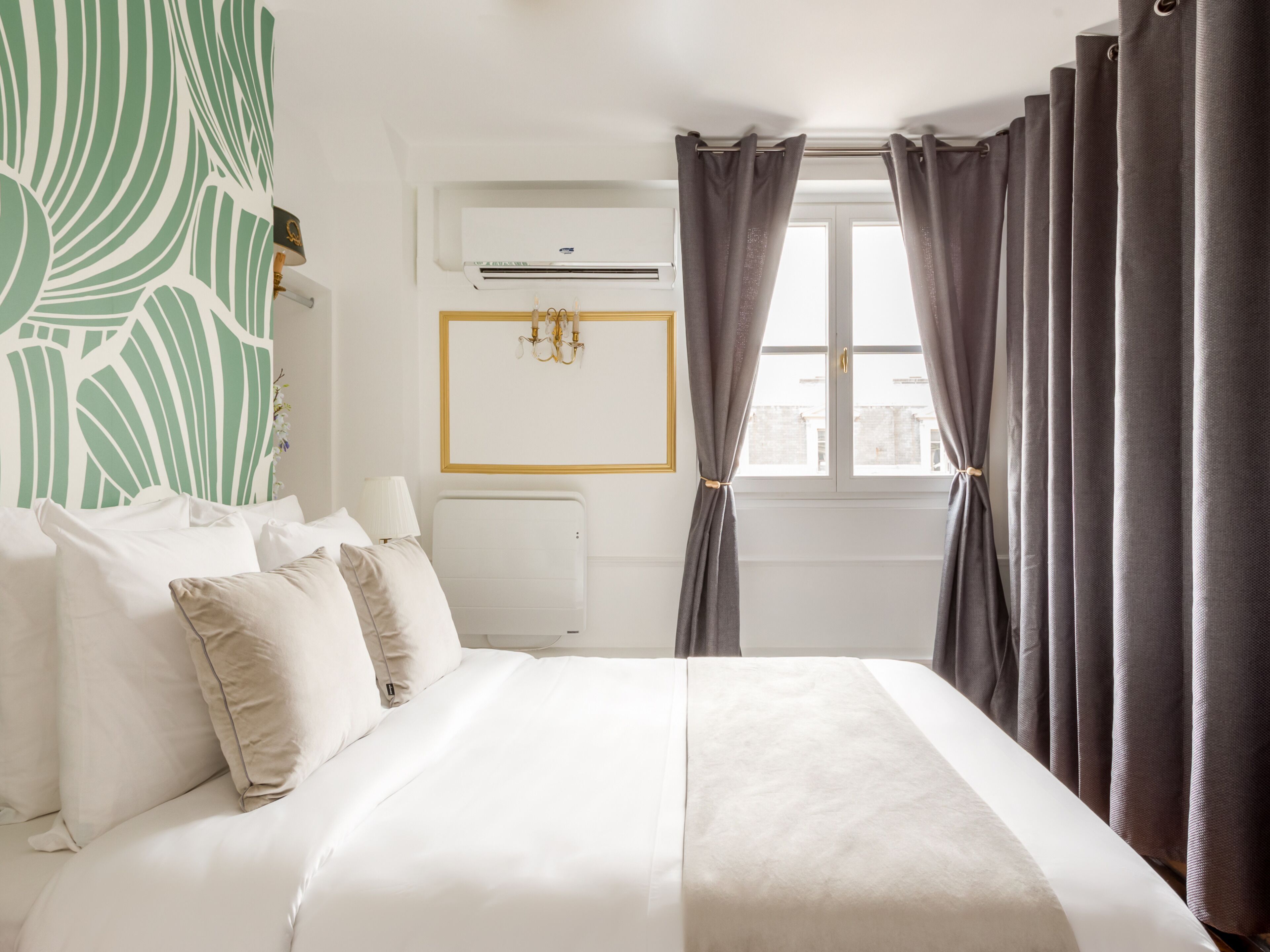 Una camera d'albergo con un letto, un condizionatore d'aria a parete, una finestra con tende e una parete decorata con un motivo a foglia verde.