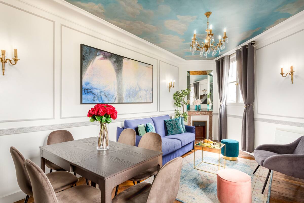 A dining area with a wooden table, cushioned chairs, a sofa, and a pink ottoman.