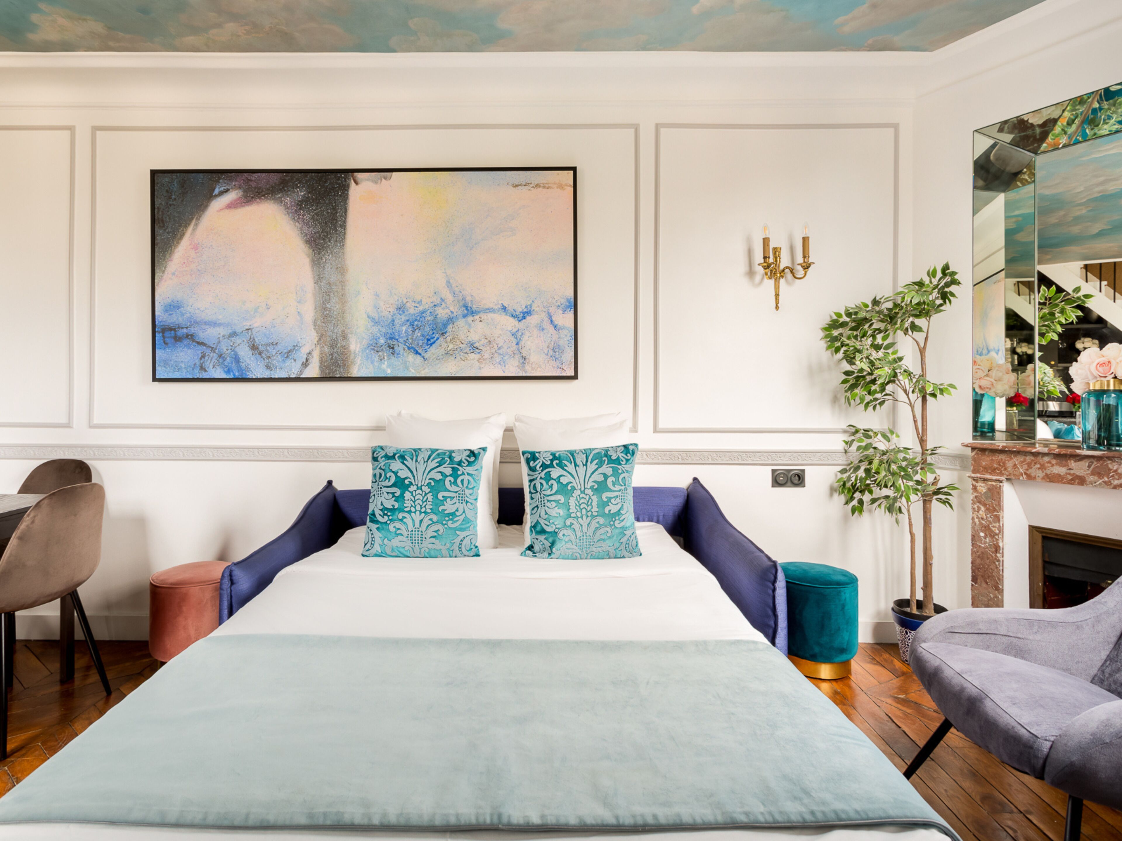 Habitación de hotel con una cama grande, un cuadro en la pared, una planta en maceta y un espejo que refleja la habitación.