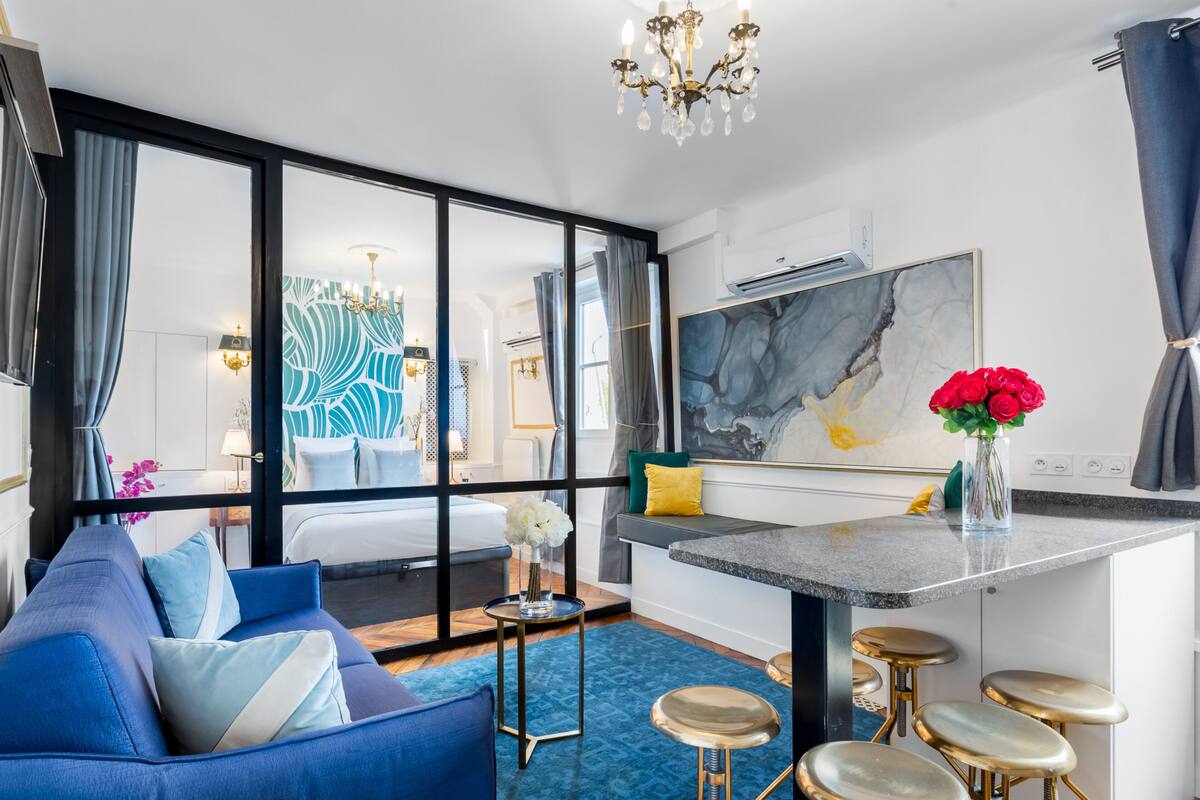 A modern hotel room with a large bed, a blue sofa, a dining area with bar stools, and a large window offering a view.