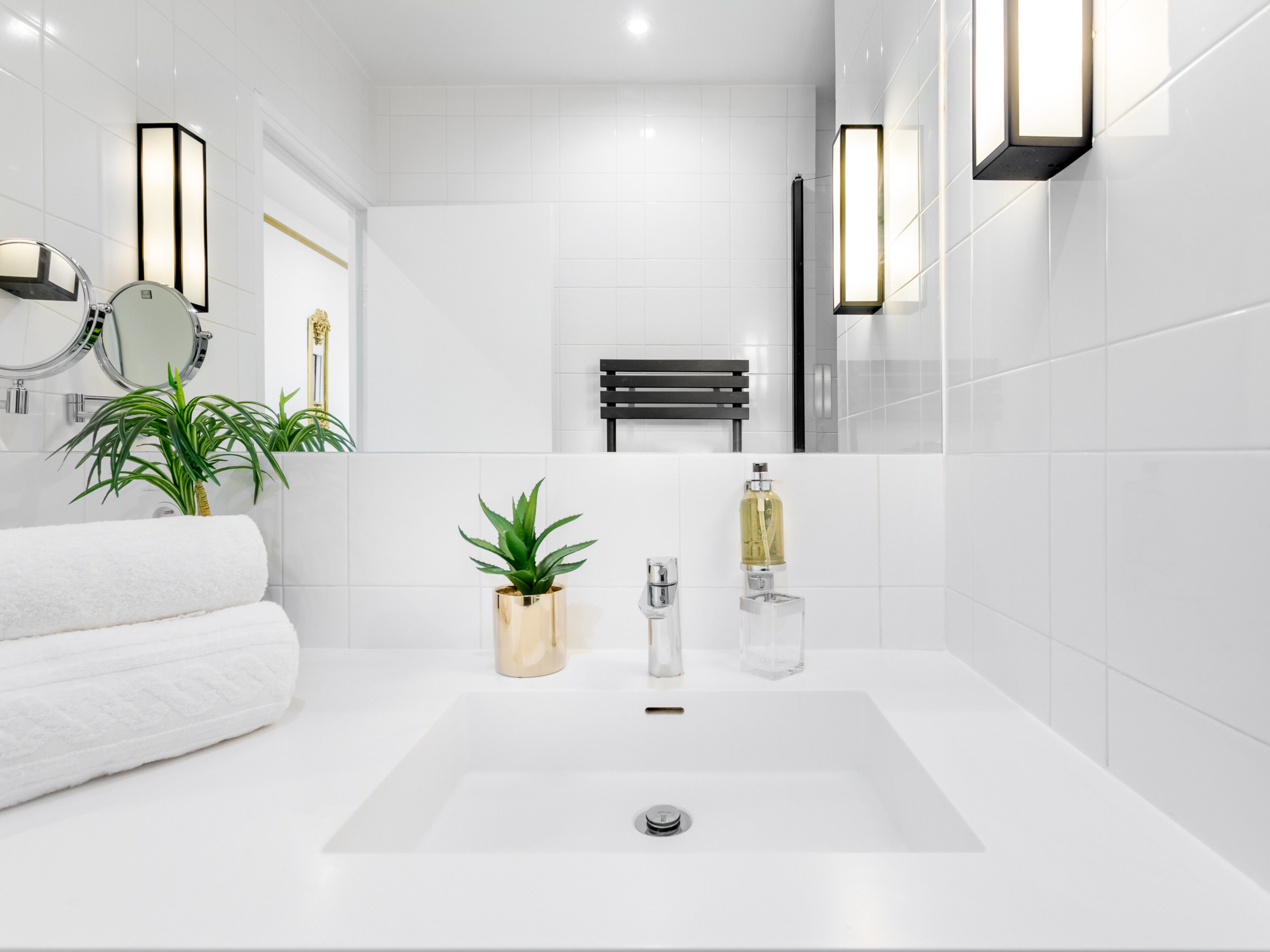 Une salle de bain moderne avec un lavabo blanc, deux plantes en pot, une serviette et un miroir.