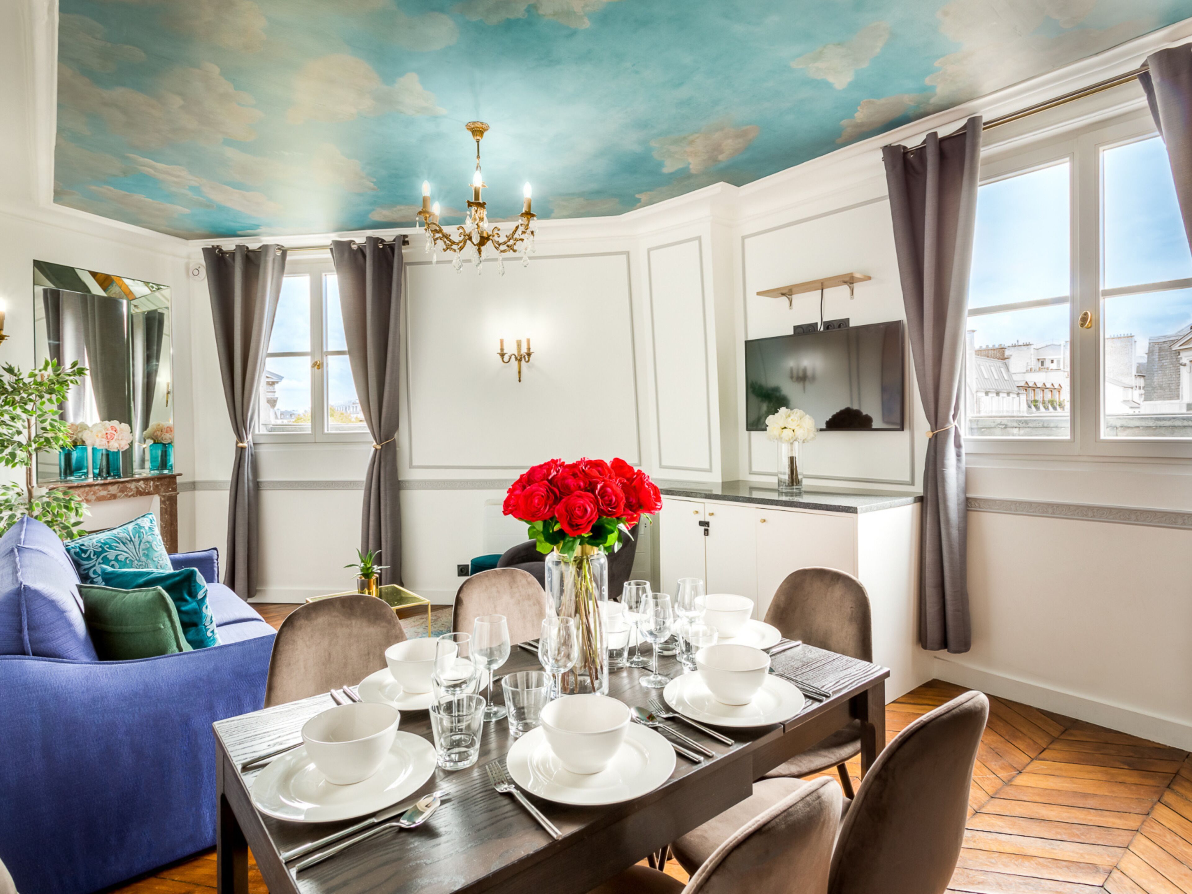 A dining room with a table set for four, a chandelier, and a view of the city through large windows.