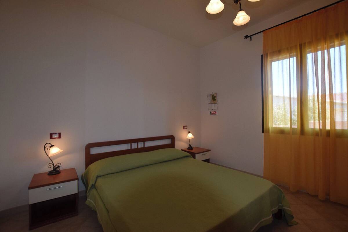 Een eenvoudige, nette slaapkamer met een enkel bed, twee nachtkastjes met lampen, een raam met gordijnen en een lichtschakelaar aan de muur.