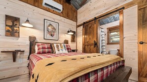 A wooden cabin bedroom with a bed, a bench, a wall-mounted shelf, a ceiling fan, and a door with a window. - Broken Arrow - Private Escape, Hot Tub & Fire Pit (Fredericksburg)