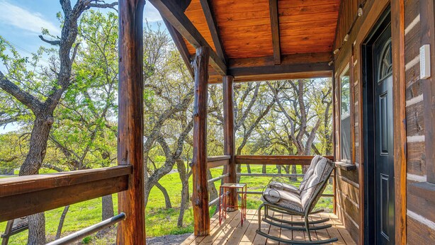 A wooden deck with a rocking chair and a small table, overlooking a grassy area with trees. - Broken Arrow - Private Escape, Hot Tub & Fire Pit (Fredericksburg)