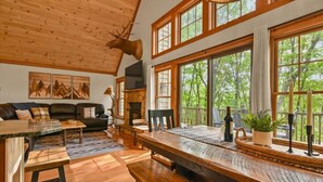Un intérieur de chalet chaleureux avec un coin repas, une cheminée et de grandes fenêtres donnant sur une forêt.