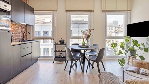 Une cuisine moderne avec un mur d’accent en briques, un coin repas avec une table ronde et des chaises, et une vue sur les bâtiments à travers de grandes fenêtres.