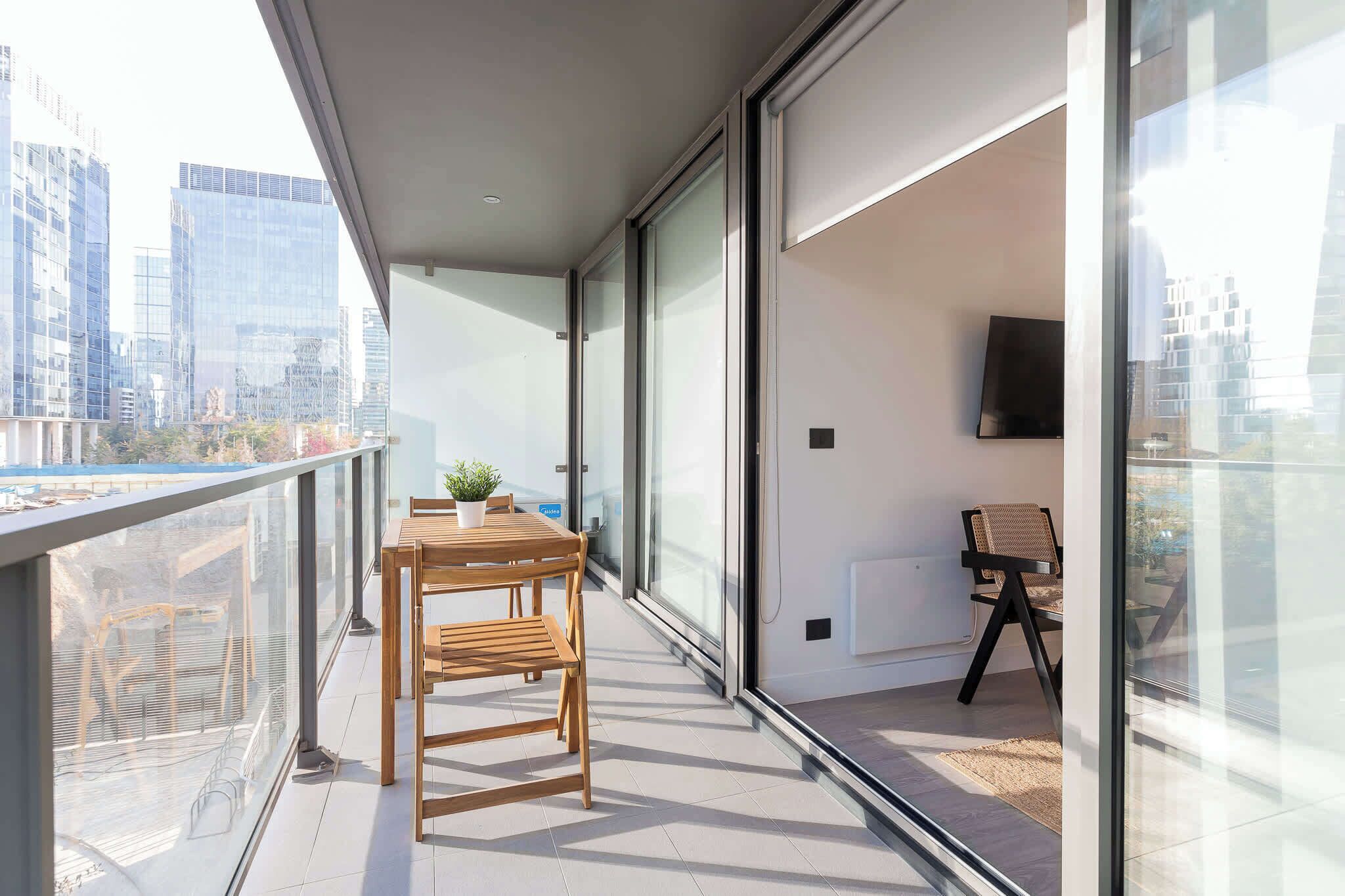 A balcony with a wooden table and chairs, a potted plant, a TV mounted on the wall, and a view of city buildings.