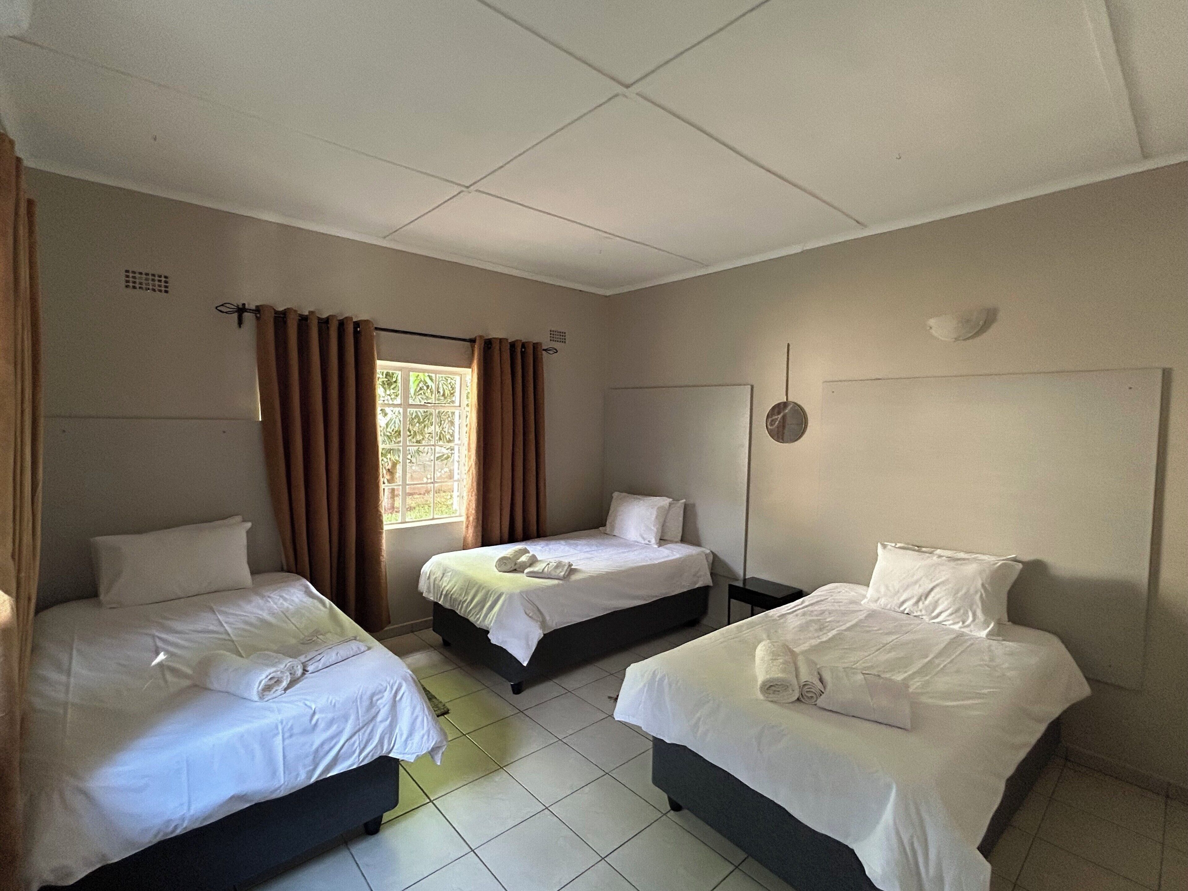 A hotel room with three single beds, a window with curtains, a wall-mounted light fixture, and a tiled floor.
