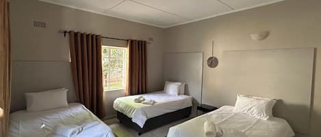 A hotel room with three single beds, a window with curtains, a wall-mounted light fixture, and a tiled floor.