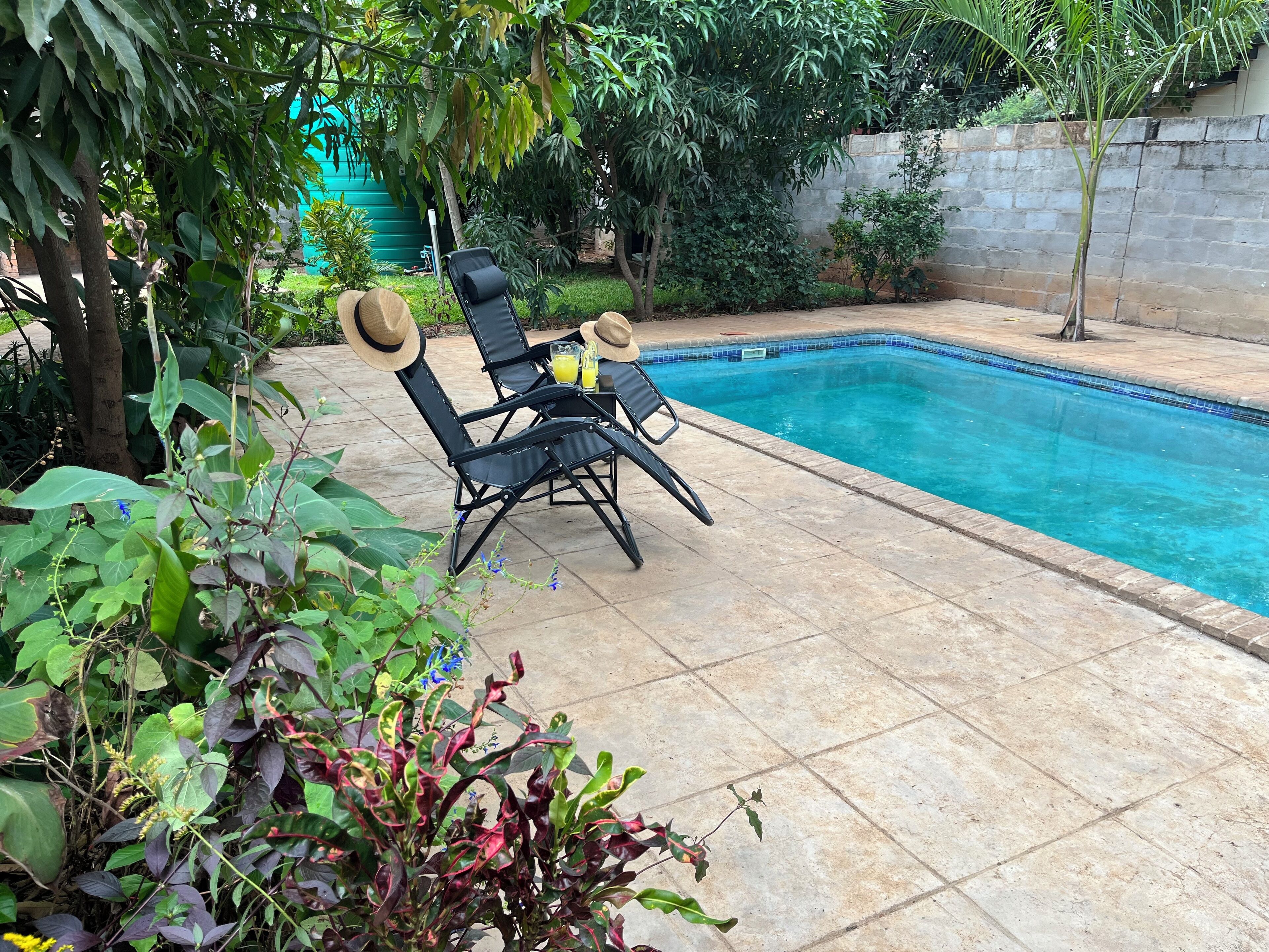Uma área de piscina com piso ladrilhado, duas cadeiras de descanso e uma mesa com uma bebida.