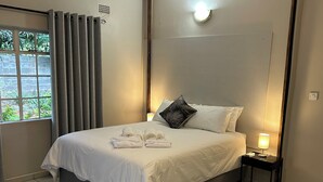 En pænt redt seng med hvide sengetøj og håndklæder, et natbord med en lampe, og udsigt til grøn vegetation gennem et vindue med gardiner.