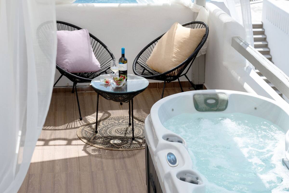A balcony with a hot tub, two wicker chairs, a table with a bottle and glasses, and a view of the sea.