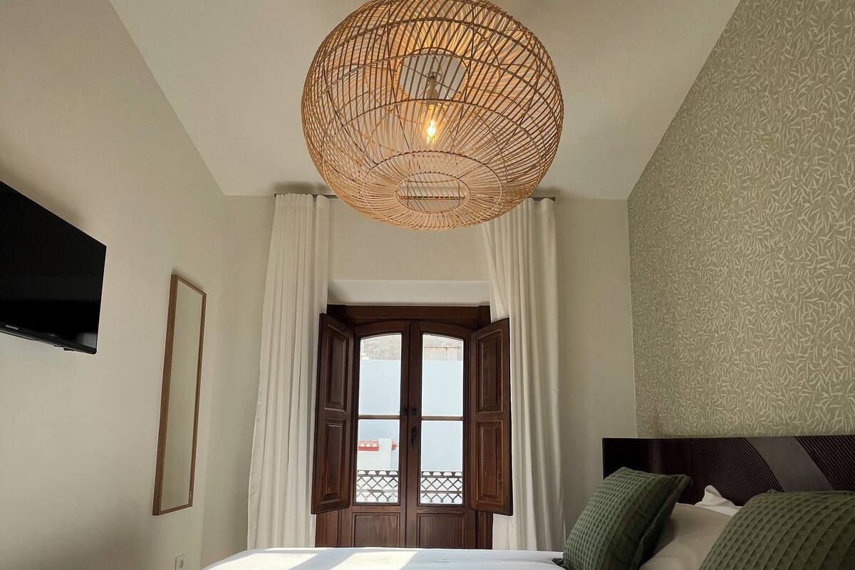 Een slaapkamer met een groot bed, een geweven plafondlamp, een aan de muur bevestigde televisie en een raam met gordijnen.