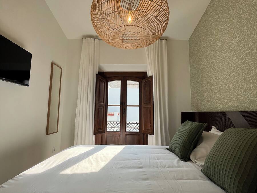 Un dormitorio con una cama grande, una lámpara de techo tejida, un televisor montado en la pared y una ventana con cortinas.