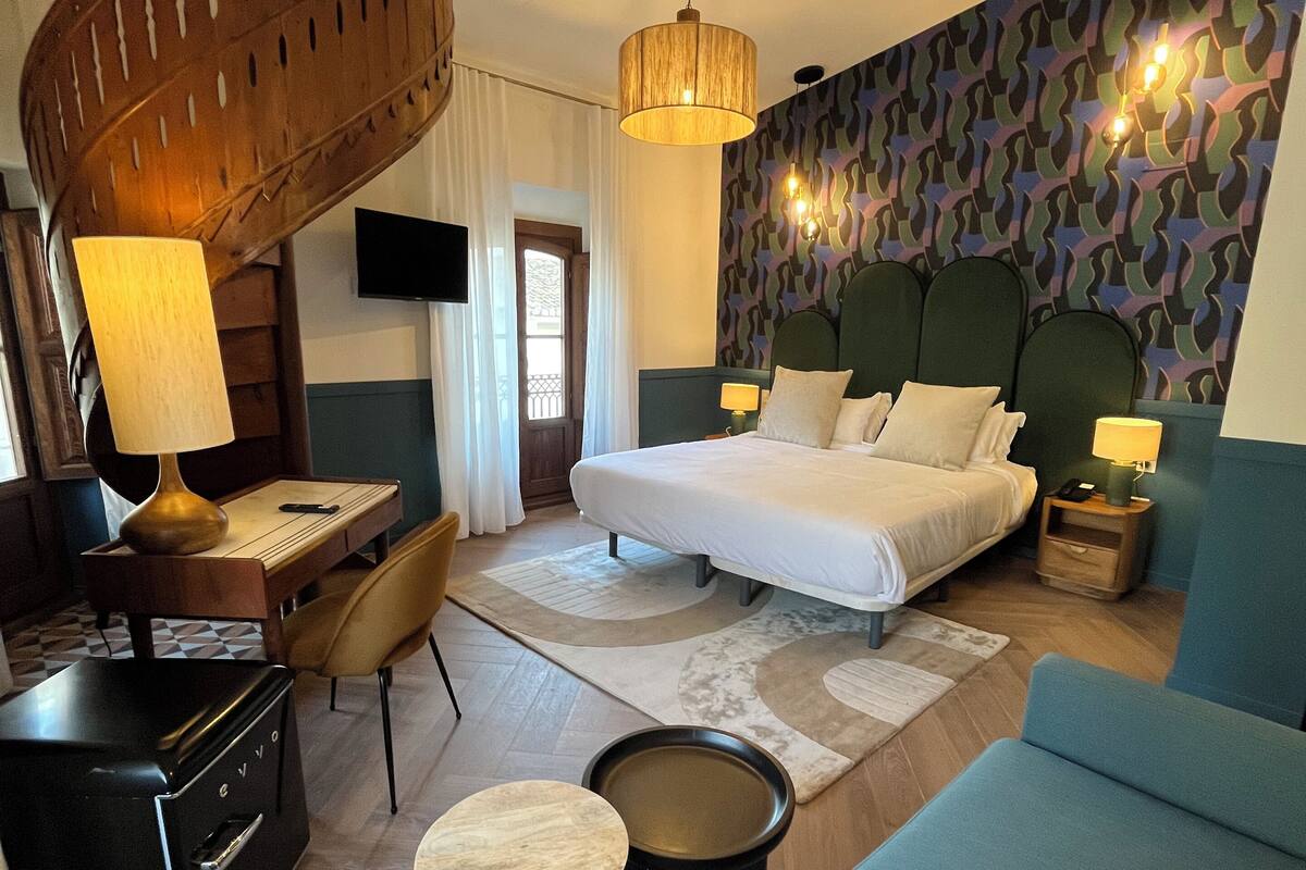 Een moderne hotelkamer met een spiltrap, een bed met veel kussens, een blauwe bank, een bureau met een lamp en een behang met een patroon.