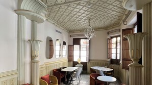 Une pièce spacieuse avec un plafond haut, des colonnes richement décorées et des carreaux de sol formant des motifs. On y trouve plusieurs tables et chaises disposées pour les repas ou les moments de convivialité.